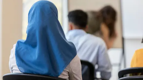 A person wearing a blue headscarf sits in a classroom, seen from behind. Photo: Getty Images.