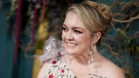 Colleen Hoover at a film premiere wearing a one-shoulder jeweled white dress, blonde hair in an updo with white feather earrings; a script tattoo is visible on her shoulder.