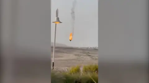 A drone is on fire about to crash to the ground. A lamppost in the foreground.