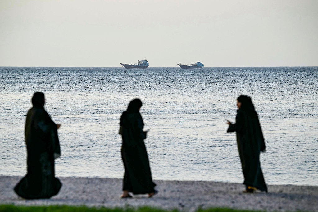 Silhouetted figures along a waterfront as cargo ships pass on the horizon, evoking oil shipping lanes near the Strait of Hormuz.